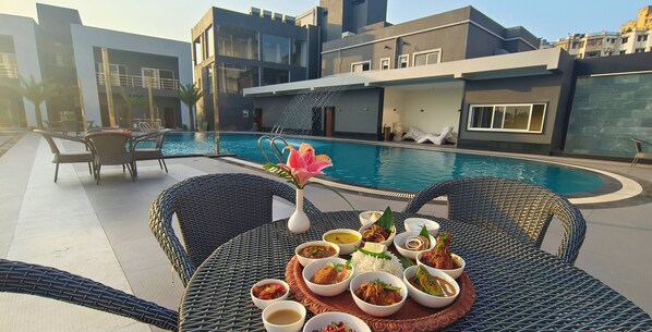 Una mesa de comedor con diversos platos, una piscina y edificios modernos al fondo.