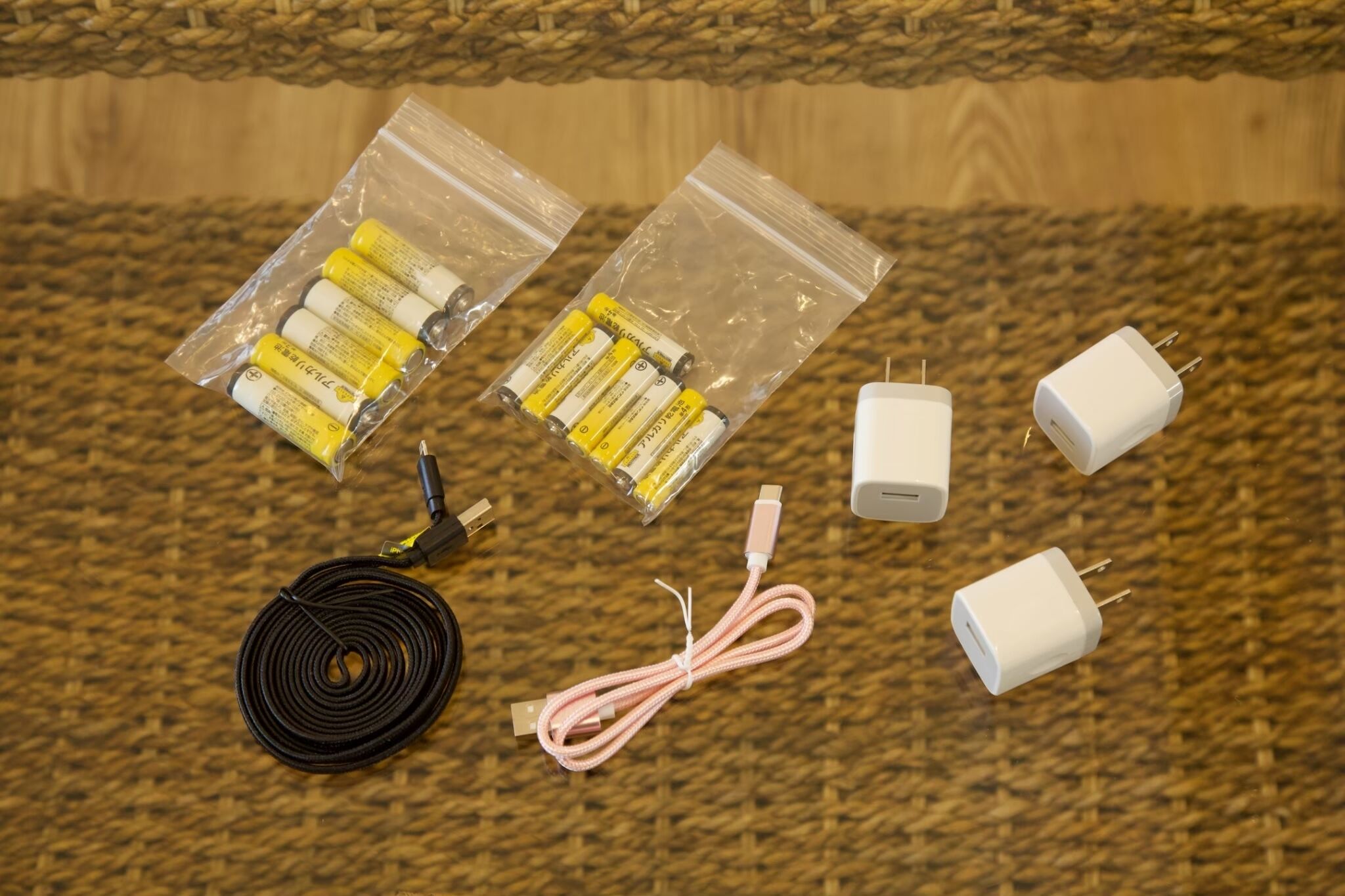 Un cavo nero arrotolato, due sacchetti di plastica trasparente con batterie gialle, due adattatori di corrente bianchi e un cavo rosa su una superficie tessuta.