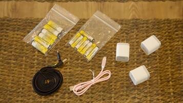 Un cavo nero arrotolato, due sacchetti di plastica trasparente con batterie gialle, due adattatori di corrente bianchi e un cavo rosa su una superficie tessuta.