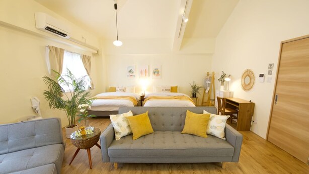 Une chambre d’hôtel avec un canapé, deux lits, une plante en pot, un plancher en bois et un ventilateur de plafond.