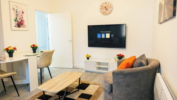 A modern living room with a flat-screen TV, a gray sofa, a wooden table, and a dining area with chairs and a small plant. - Central Cardiff Cozy 2BR Near Stadium Shops Uni (Cardiff)