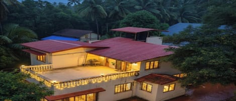 A two-story house with a red roof, surrounded by lush greenery and palm trees.