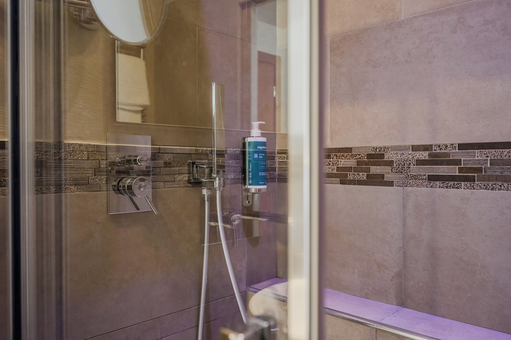 A glass-enclosed shower with a handheld showerhead, a wall-mounted faucet, and a soap dispenser.