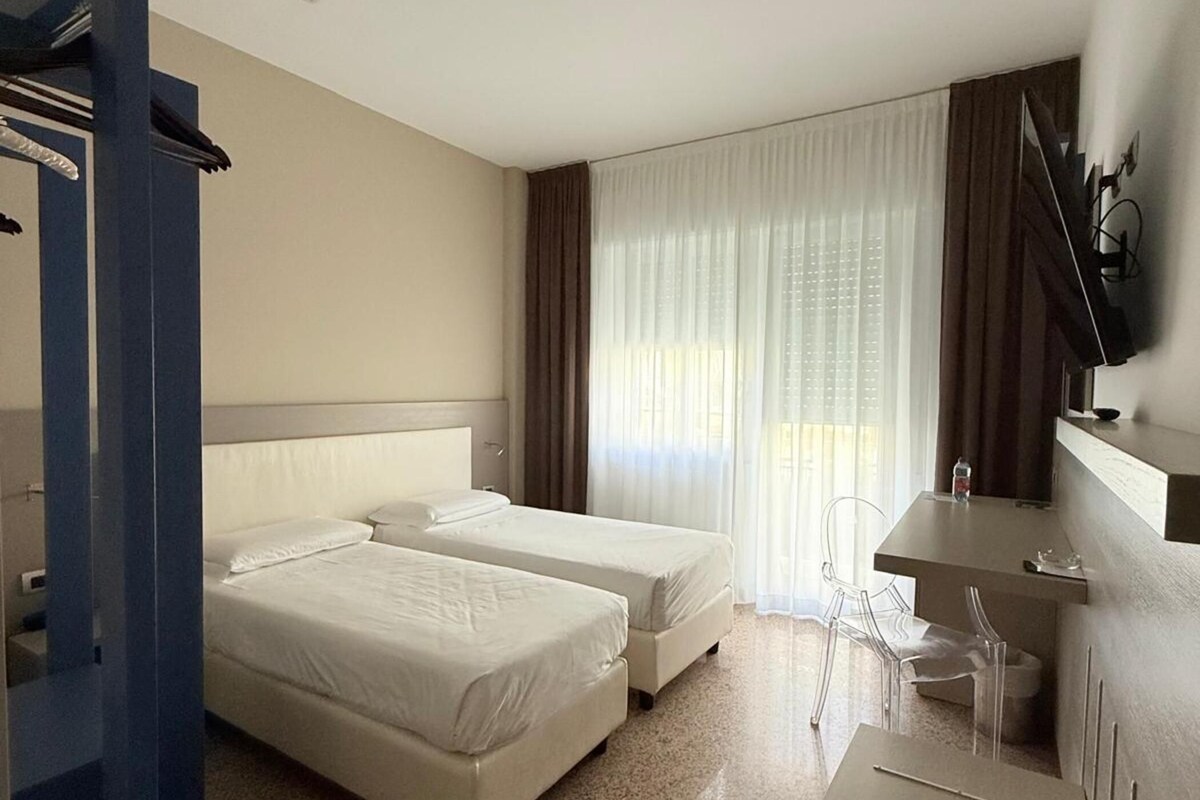 A hotel room with two beds, a desk, a chair, and a large window with curtains.