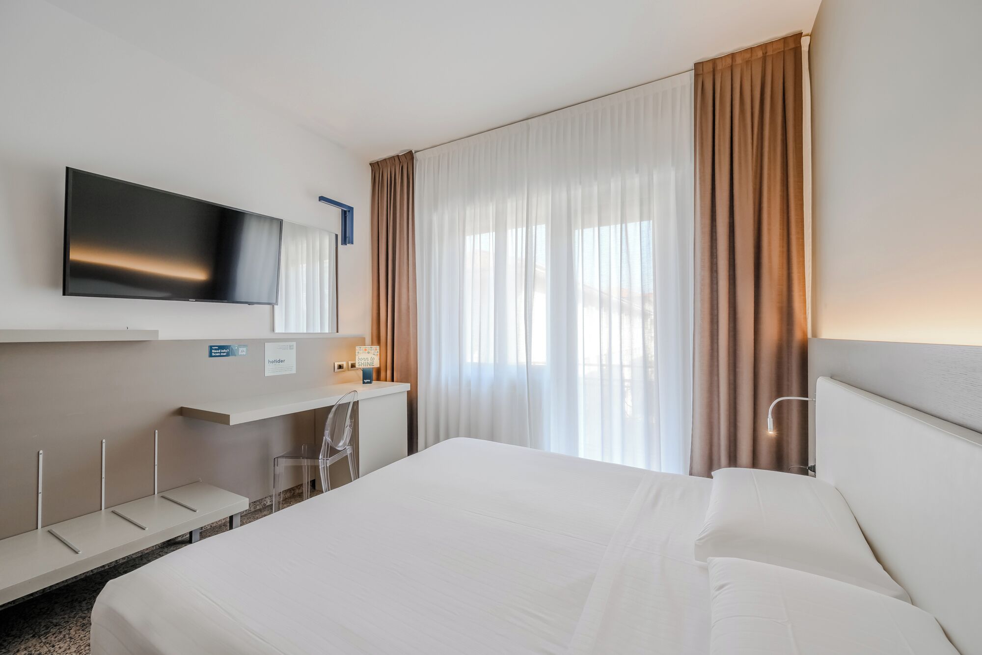 Habitación de hotel con cama, televisor montado en la pared, escritorio con silla y ventana con cortinas.