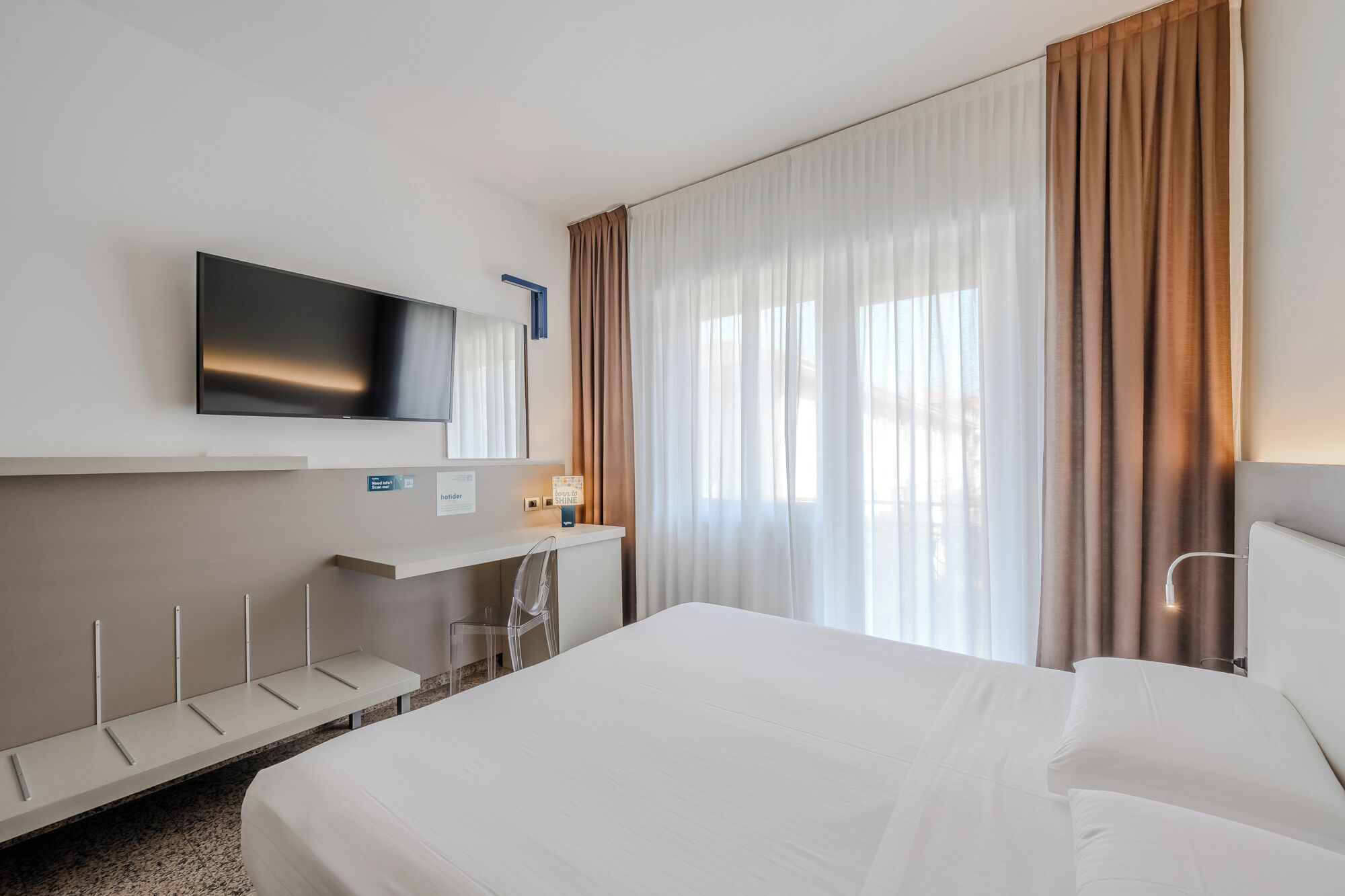 Quarto de hotel com uma cama, televisão na parede, escrivaninha com cadeira e janela com cortinas.