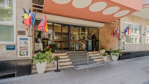 Entrada de hotel con puerta de cristal, plantas en macetas y varias banderas.
