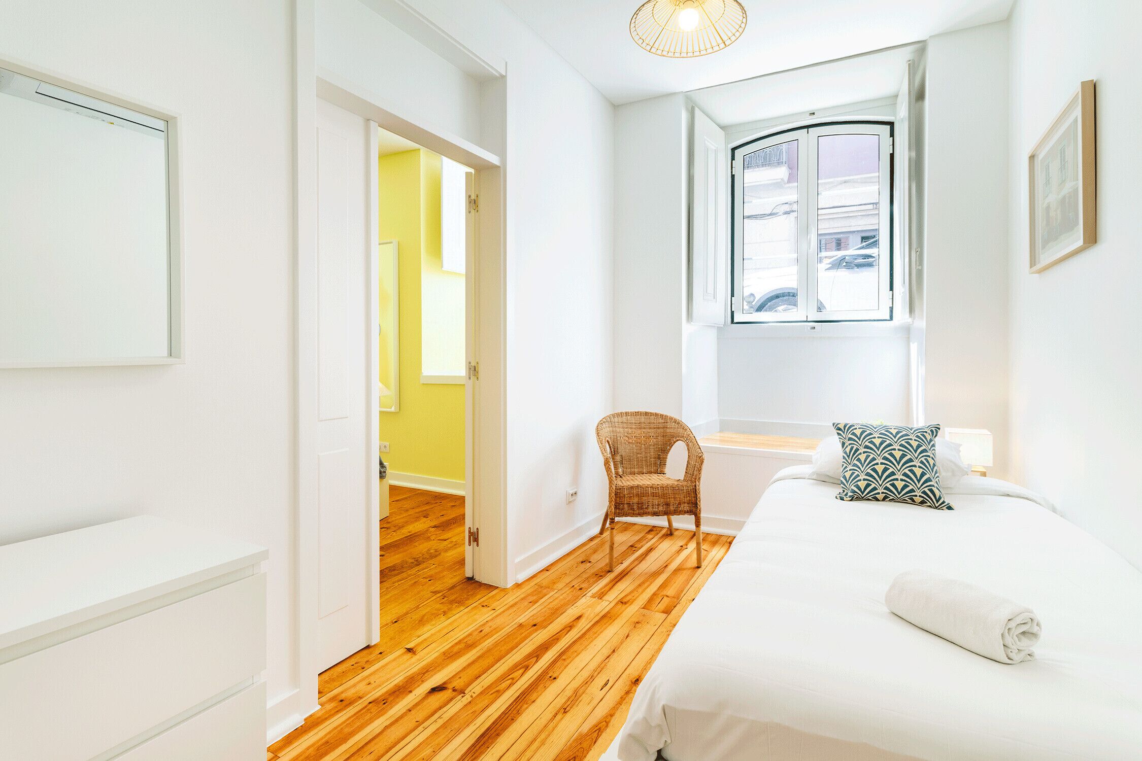 Een slaapkamer met een houten vloer, een bed met witte beddengoed, een rieten stoel, een raam met uitzicht en een deur die leidt naar een andere kamer.
