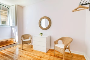 Una habitación con piso de madera, un tocador blanco, dos sillas de mimbre, un espejo redondo y una ventana con vistas a edificios.