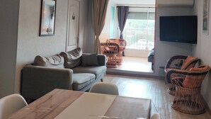 Un salon comprenant un canapé, une table à manger, des chaises en osier, un téléviseur à écran plat et une grande fenêtre.