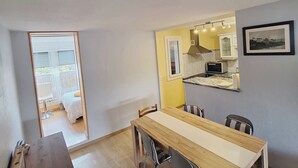 Un coin repas avec une table et des chaises en bois, une cuisine visible à travers une porte ouverte et un balcon avec un lit.