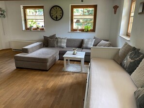 A living room with a sectional sofa, a small table, and two windows with plants.