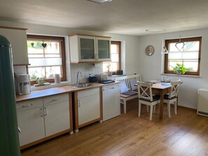 A compact kitchen with wooden cabinets, a built-in dishwasher, a dining table with chairs, and a window with a plant.