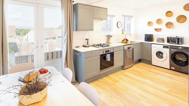 A modern kitchen with grey cabinets, a built-in oven, a washing machine, and a dining area with a table and chairs. - Contractor Home Parking & Garden – Near Avonmouth (Bristol)