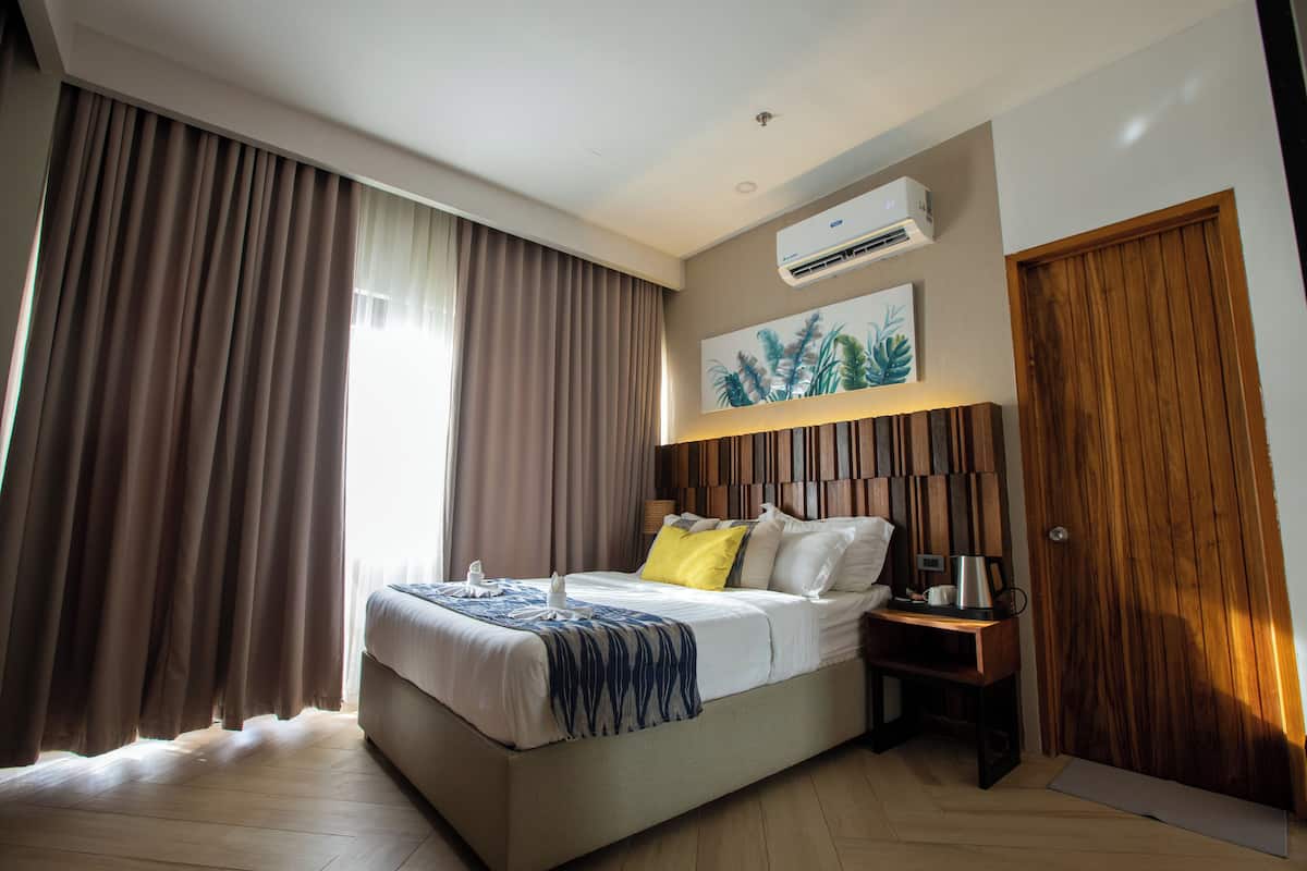 A hotel room with a bed, a nightstand, a coffee maker, a wooden door, and an air conditioning unit.