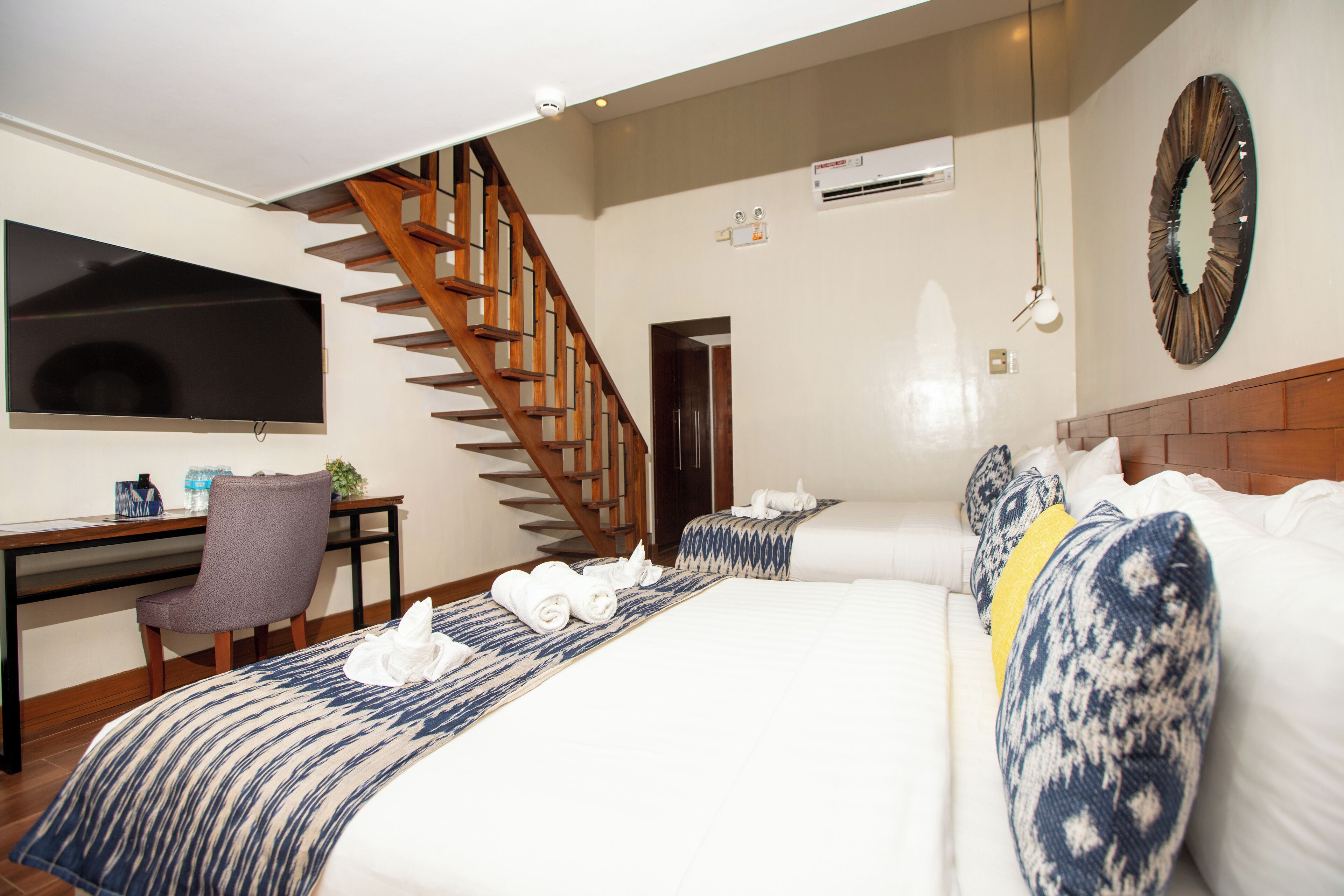 A hotel room with a large bed, a desk with a chair, a TV, a wooden staircase, and an air conditioning unit.