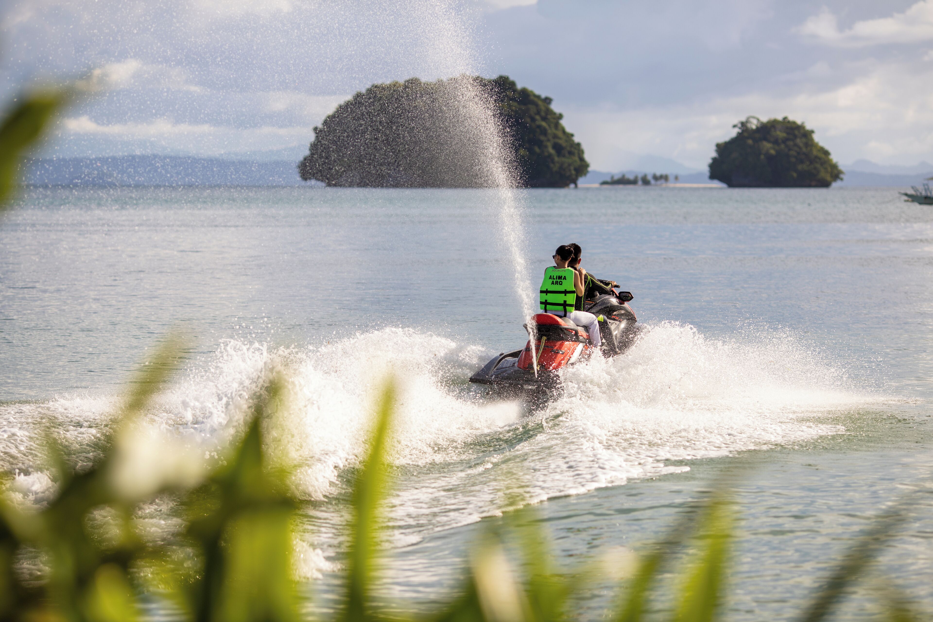 Un jet ski avec une personne portant un gilet de sauvetage au premier plan, de l’eau qui éclabousse tout autour, et deux îles en arrière-plan.