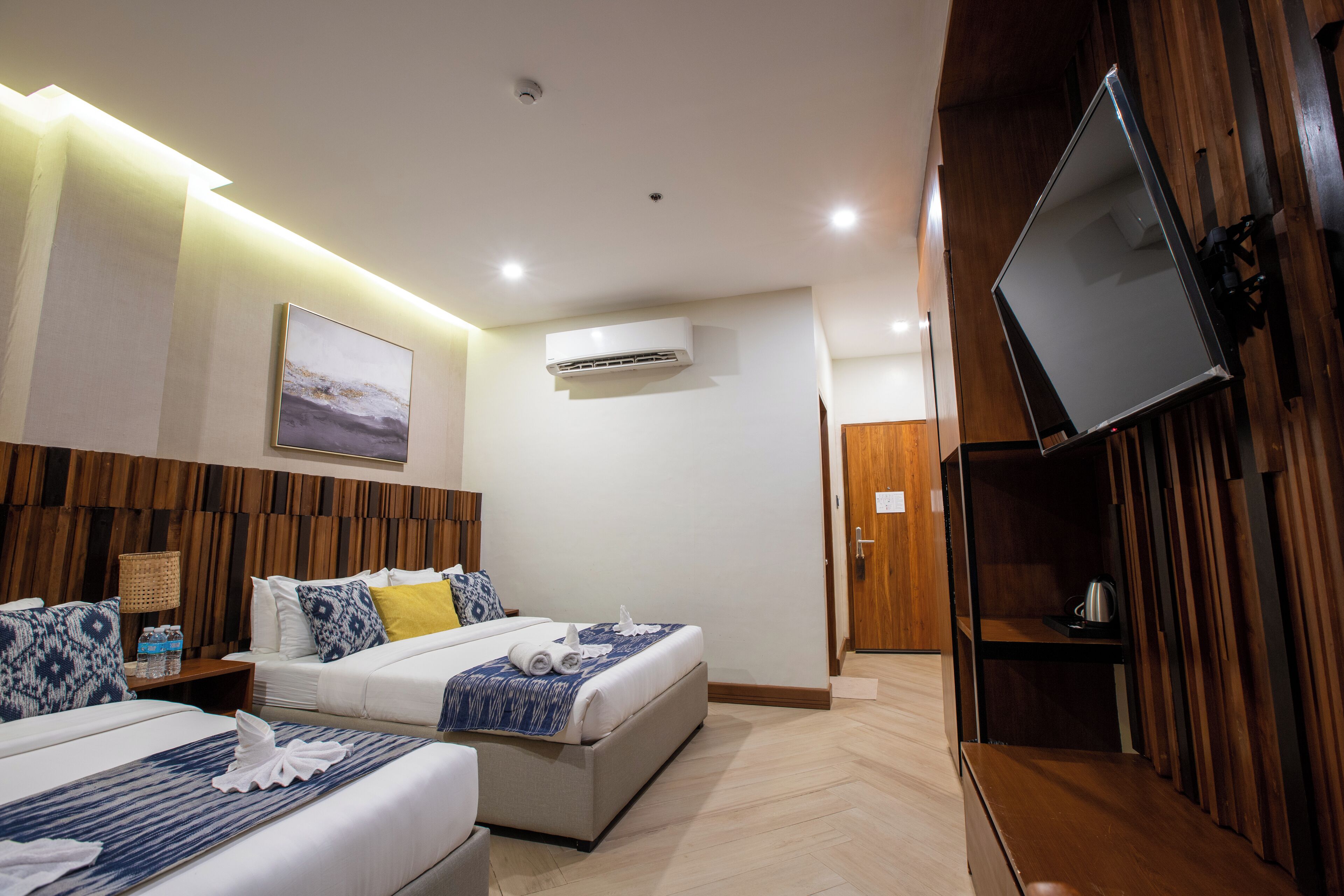 A hotel room with two beds, a flat-screen TV mounted on the wall, an air conditioning unit, and a wooden panel wall.