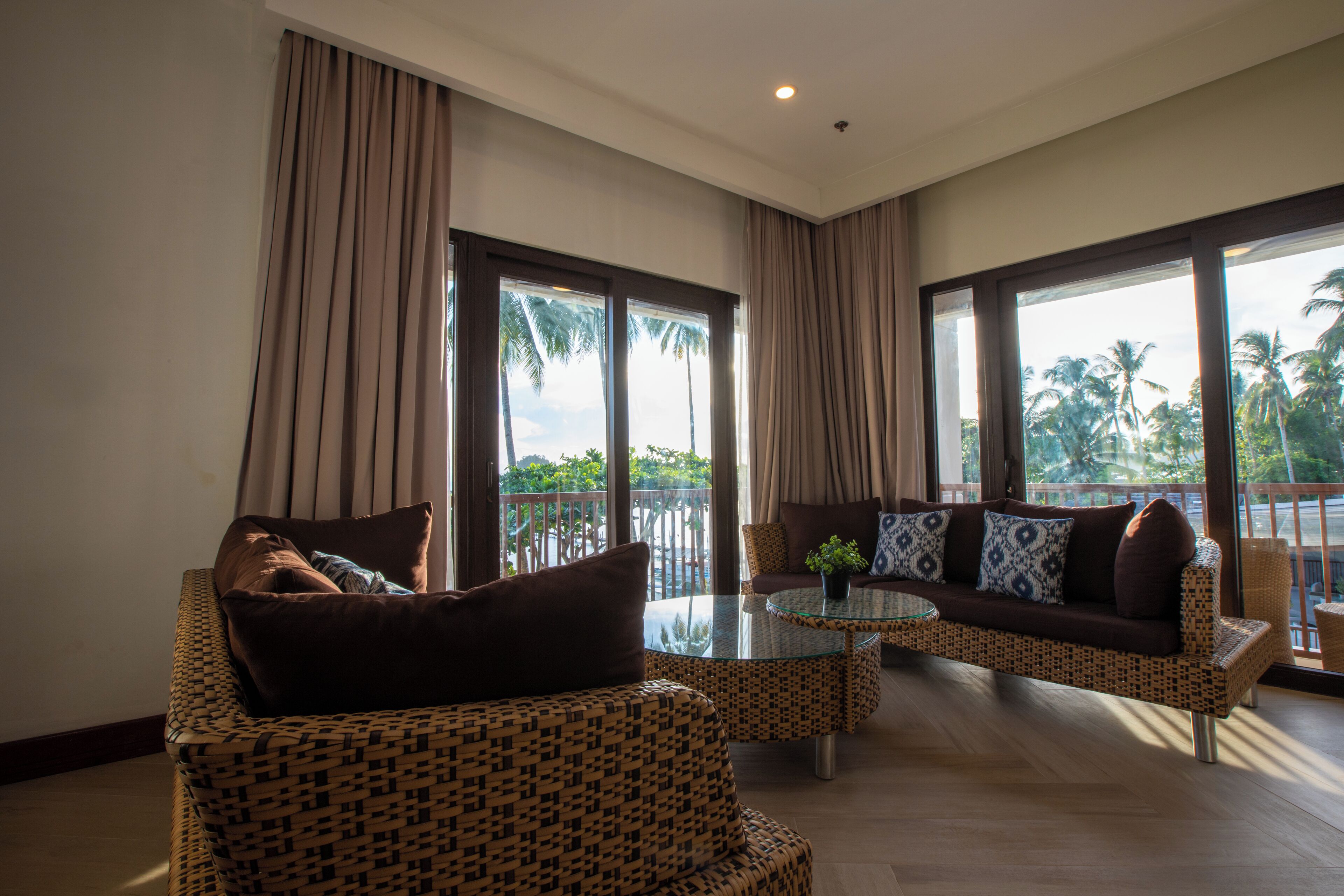 A spacious living area with a sofa, armchairs, and a glass coffee table. Large windows provide a view of palm trees and a balcony.