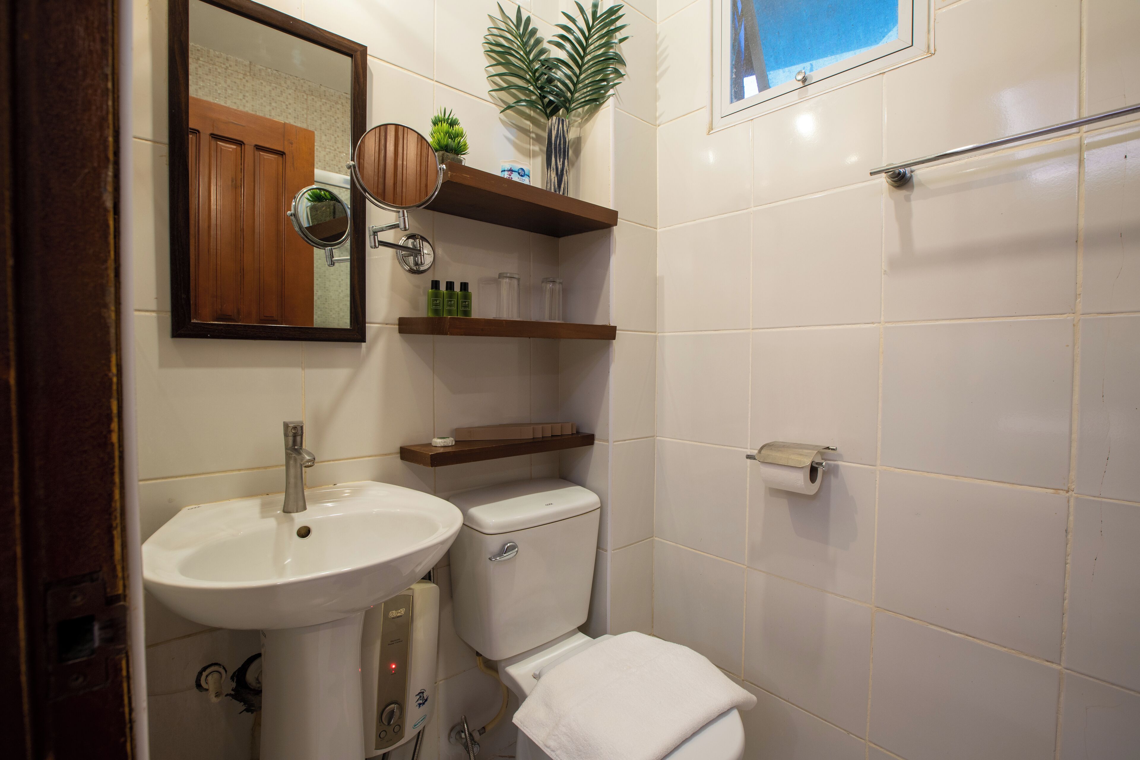Un bagno con lavandino, water e specchio bianchi. Sono presenti mensole in legno sopra il water e una piccola pianta sulla mensola più in alto.