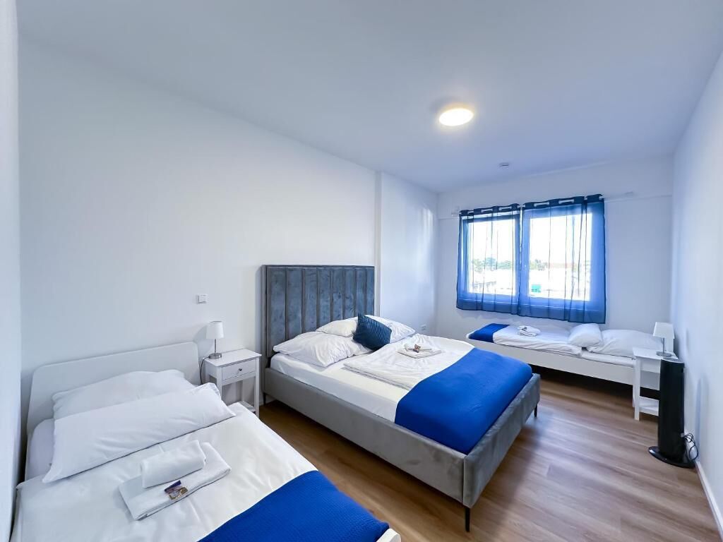 Habitación de hotel con dos camas, cabecero de madera, mesita de noche con lámpara y ventana con cortinas.