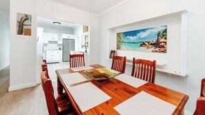 A dining area with a wooden table, white placemats, and wooden chairs. There is a painting of a beach scene on the wall. - The Waratah Escape Nowra (Nowra)