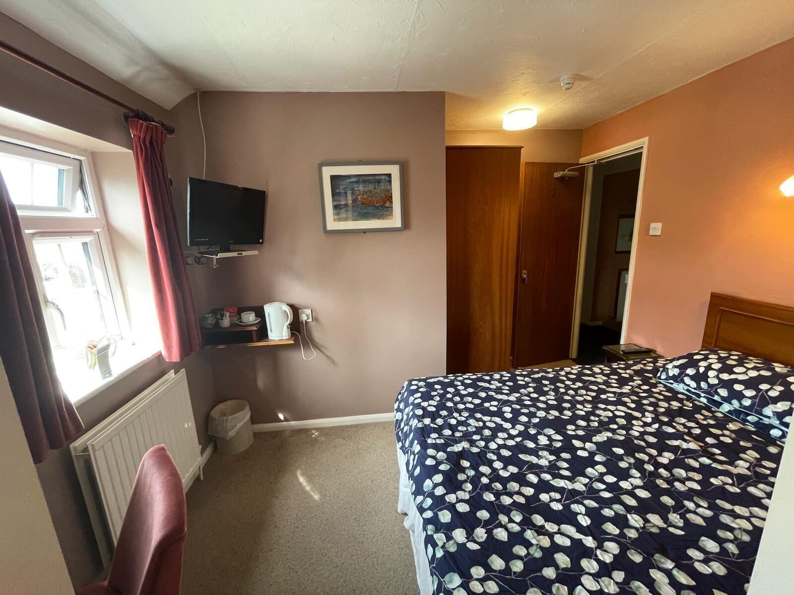 Ein Hotelzimmer mit Bett, Fernseher, einem gerahmten Bild an der Wand, einer Heizung und einem Fenster mit Vorhängen.