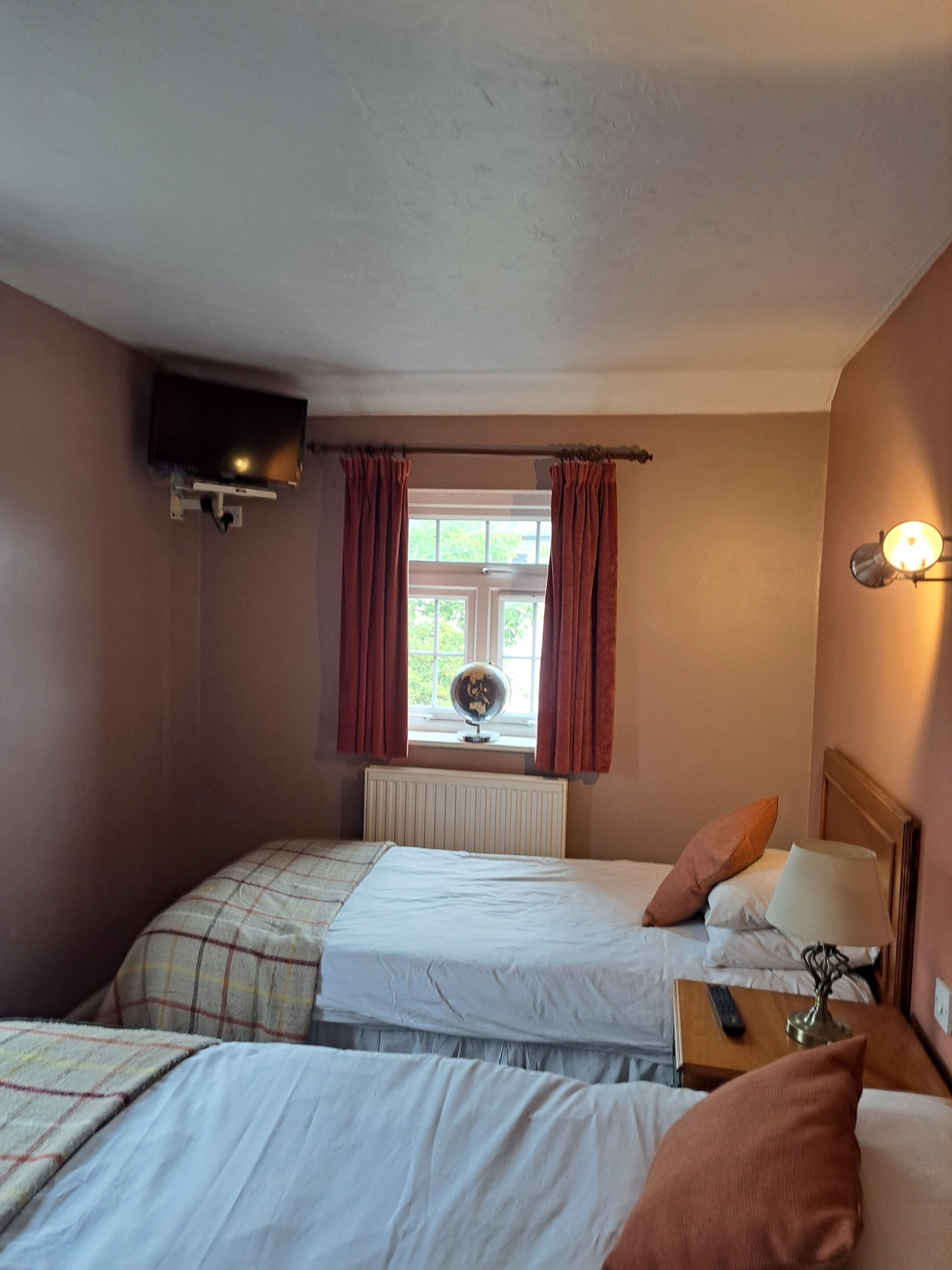 A hotel room with two beds, a television mounted on the wall, a window with curtains, and a bedside table with a lamp.