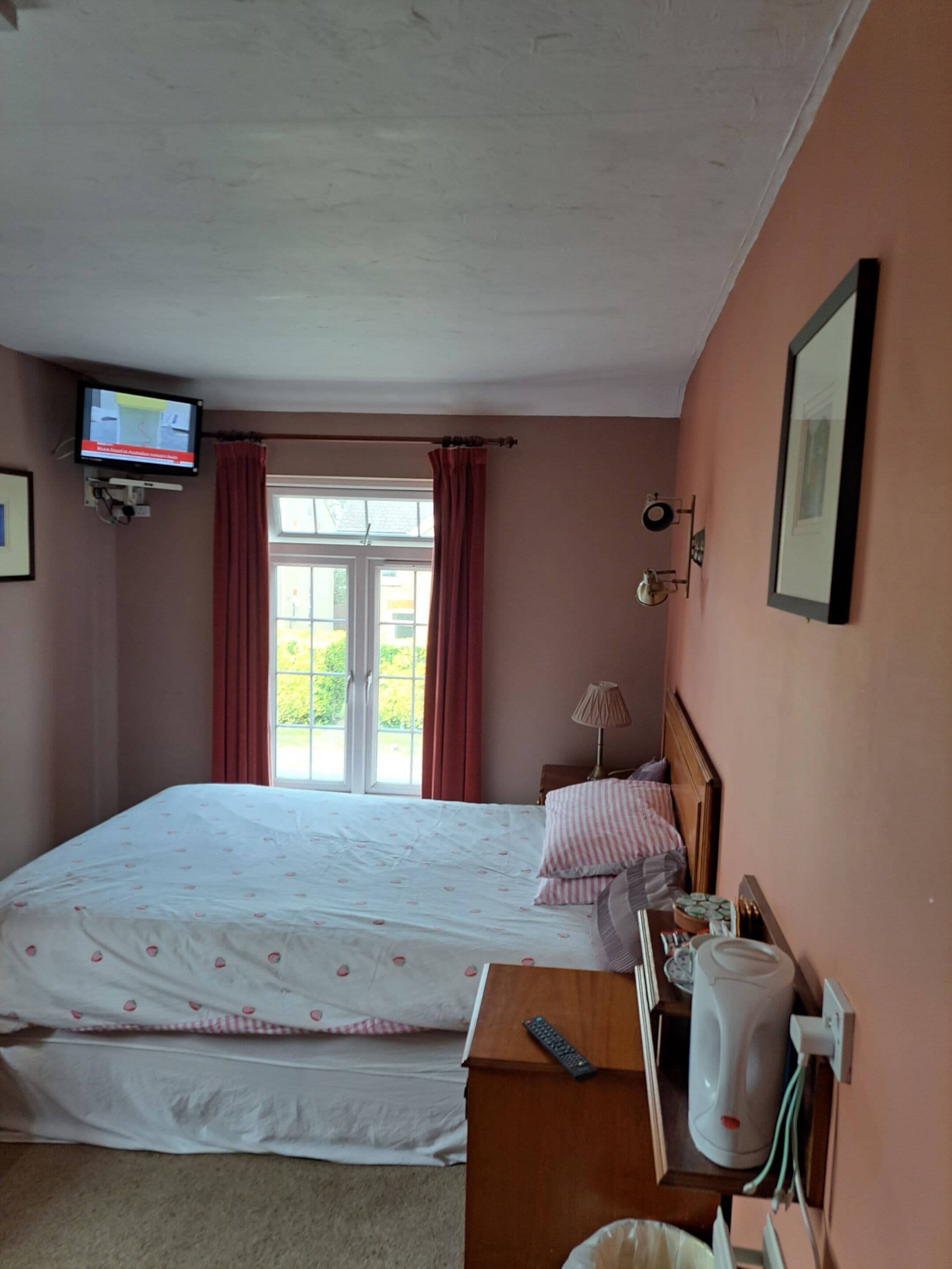 A bedroom with a bed, a television mounted on the wall, a window with curtains, and a bedside table with a kettle and a remote control.