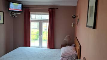 A bedroom with a bed, a television mounted on the wall, a window with curtains, and a bedside table with a kettle and a remote control.