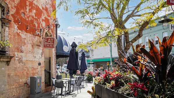 A street view with a Thai eatery sign, outdoor seating, and tropical plants.