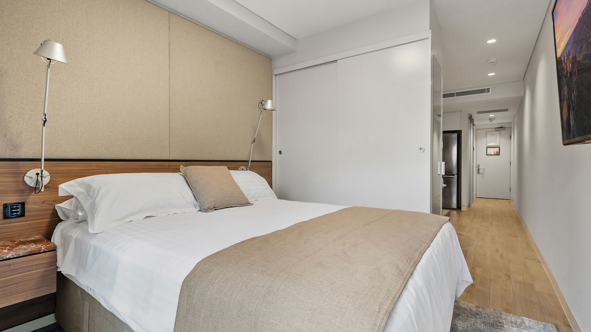 A hotel room with a bed, bedside lamps, a wooden headboard, a nightstand, and a door leading to a hallway.