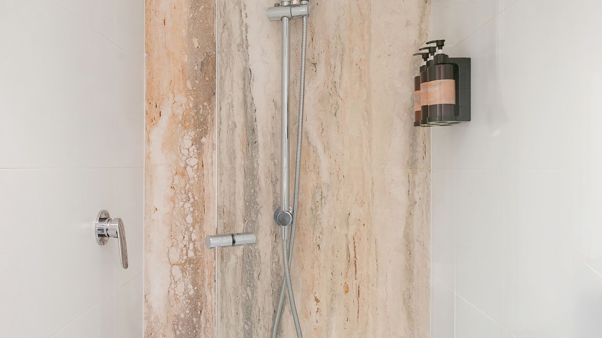 A bathroom with a marble wall, a shower with a handheld showerhead, and a soap dispenser on the wall.