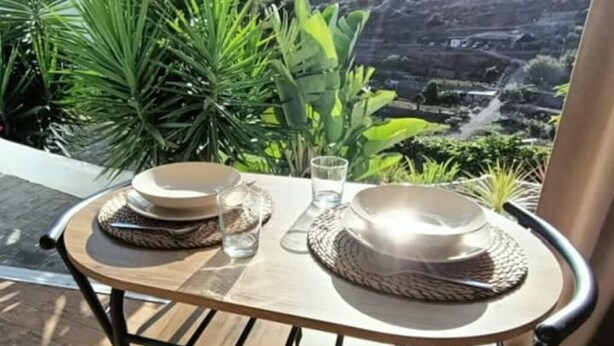 A round table with a woven placemat, a glass of water, and a bowl on a balcony with a view of a valley. - Estudio Nuevo con Jardín Privado en Pleno Valle (La Laguna)