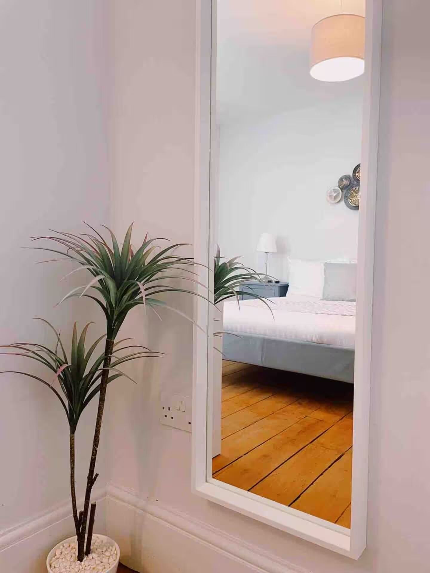 Una habitación con una cama, una planta en maceta y un espejo que refleja una lámpara y adornos de pared.