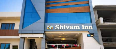 A modern hotel named Shivam Inn with a prominent blue and brown facade, a visible entrance with a staircase, and a car parked in front.
