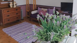Een gezellige kamer met een houten dressoir, een bed met paars beddengoed, een staande lamp en een groot raam.