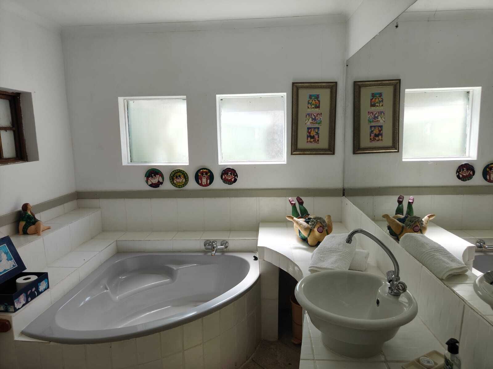 Casa de banho com banheira, pia e um espelho grande. Há quadros nas paredes e um pequeno armário com artigos de higiene pessoal.
