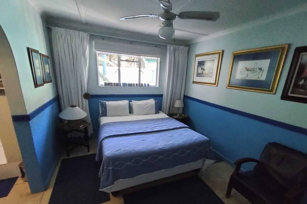 Une chambre à coucher avec un lit, un ventilateur de plafond, une fenêtre avec des rideaux, une chaise et des tableaux encadrés accrochés au mur.