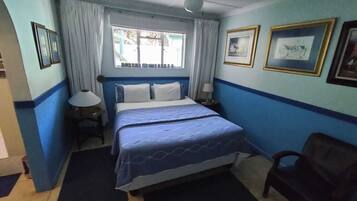 Un dormitorio con cama, ventilador de techo, ventana con cortinas, una silla y cuadros en la pared.