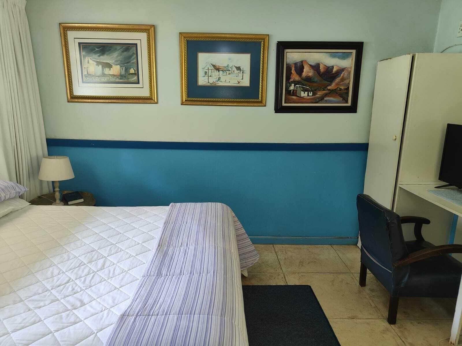 Quarto de hotel com cama, escrivaninha, cadeira e três quadros na parede.