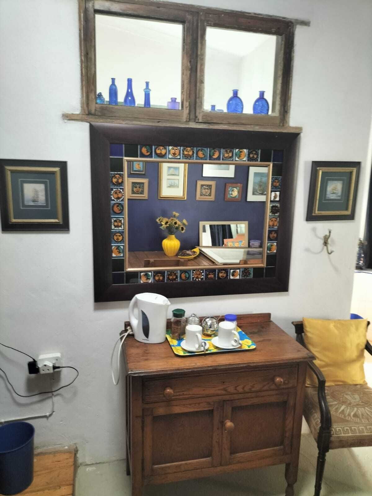 Une pièce avec un meuble en bois, une petite table sur laquelle se trouve un plateau avec des tasses, une fenêtre ornée de bouteilles bleues et des tableaux encadrés accrochés au mur.