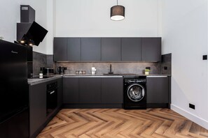 A modern kitchen with dark cabinetry, a built-in washing machine, and a herringbone wood floor. - Weaver Suite (Ayr)