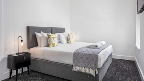Un lit bien fait, avec une tête de lit grise, du linge de lit blanc et un plaid à motifs. Une petite table de chevet avec une lampe est visible sur la gauche.