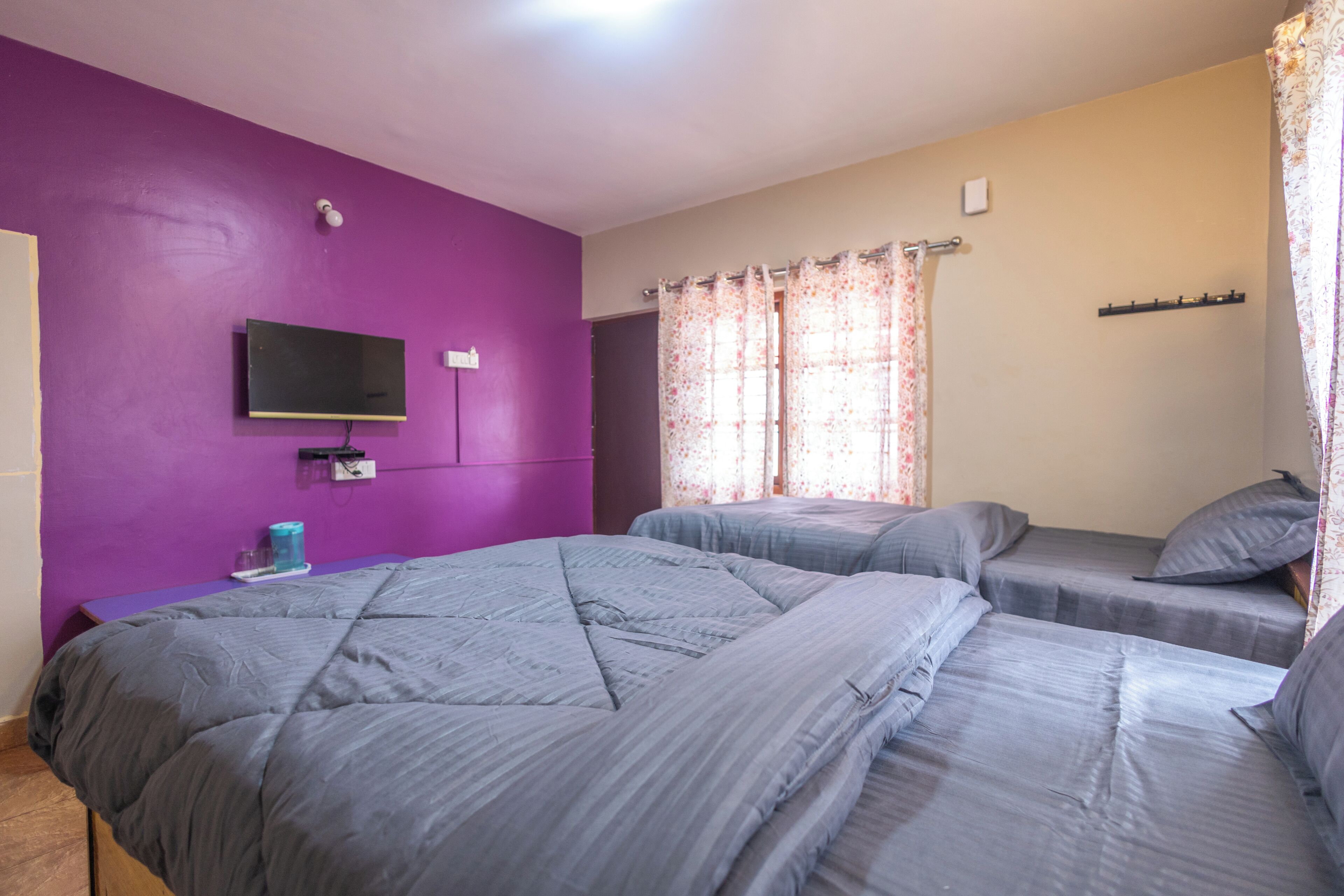Une chambre avec deux lits, un téléviseur fixé au mur et une fenêtre ornée de rideaux à motifs floraux.