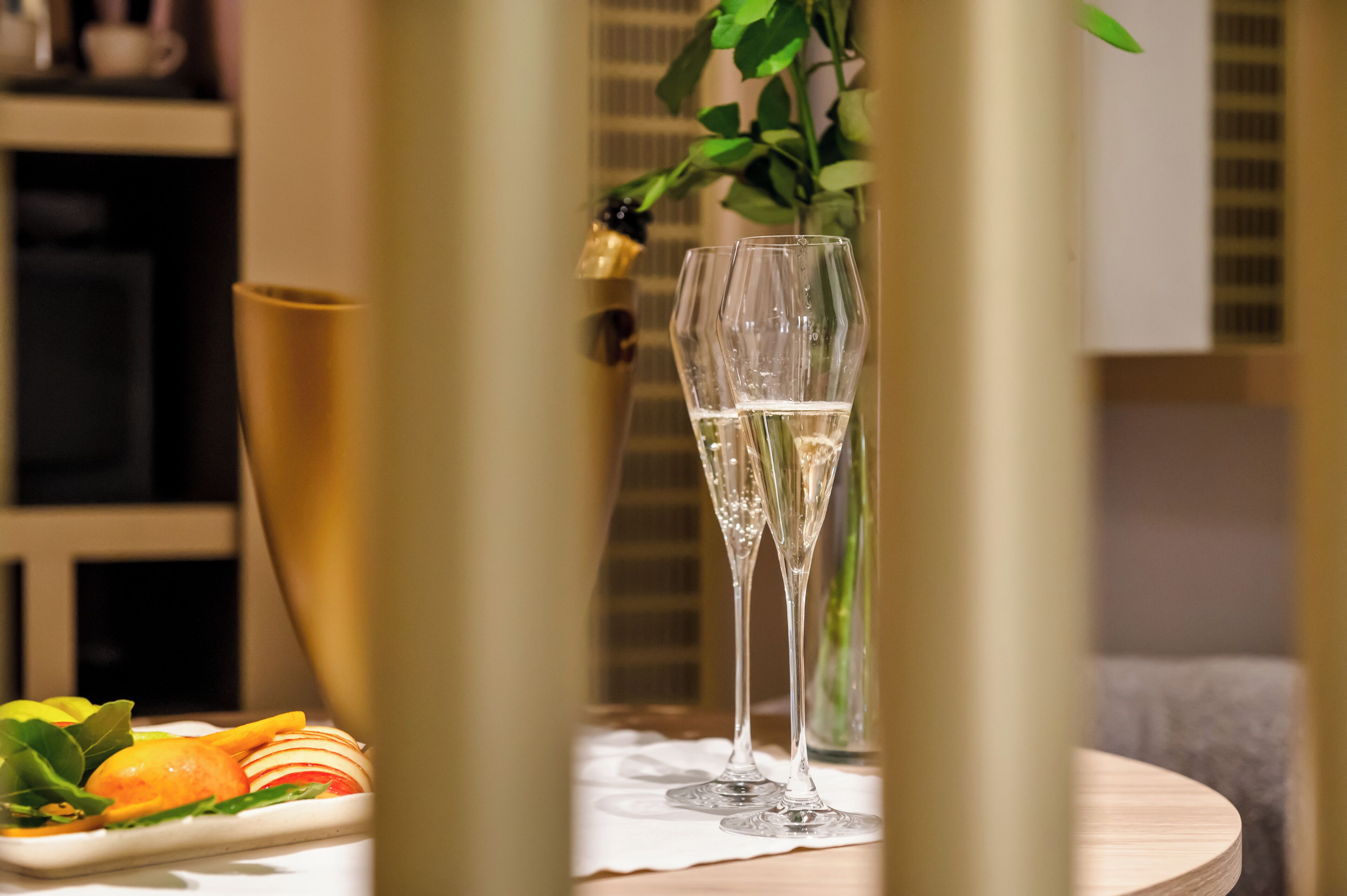 A table with two champagne flutes, a vase with green leaves, and a plate with sliced fruits.