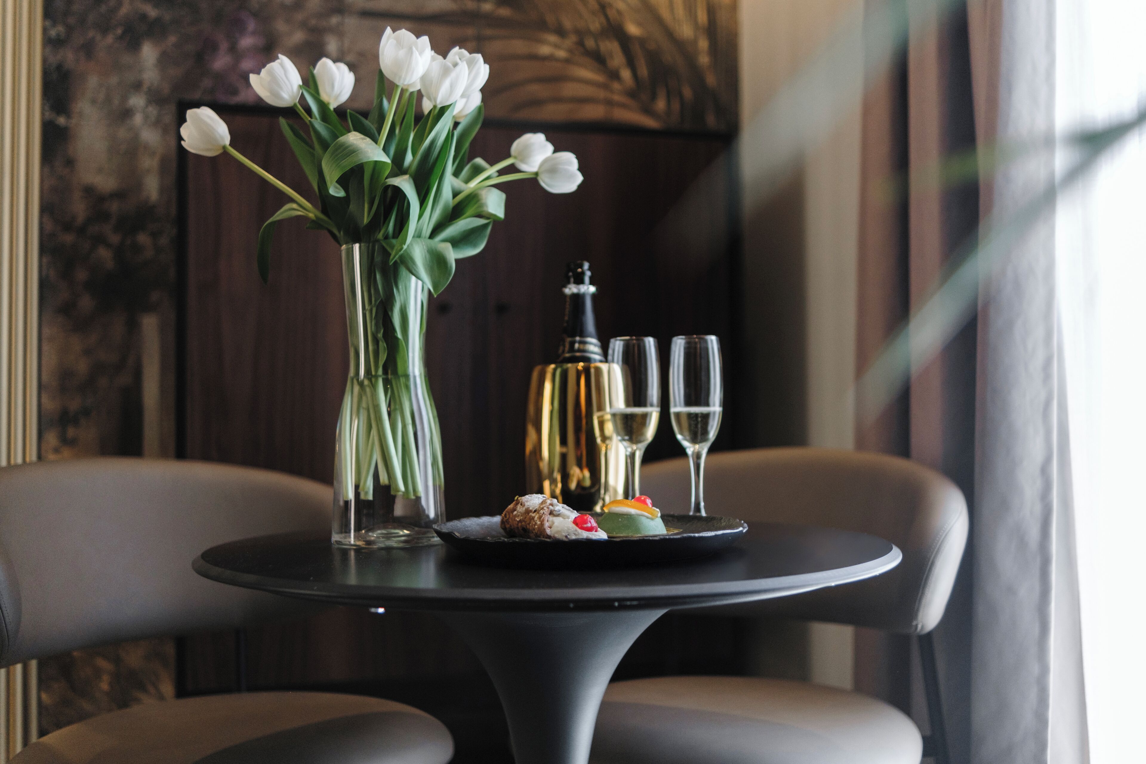 Une table avec un vase de tulipes blanches, une bouteille, deux verres et une assiette contenant un dessert.