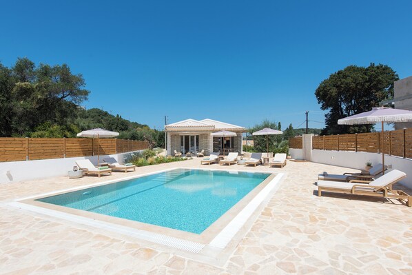 A swimming pool with lounge chairs and umbrellas, surrounded by stone flooring and wooden fences.