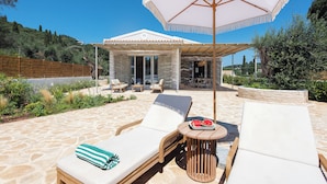 Un espace de détente extérieur, baigné de soleil, avec un sol en pierre, deux fauteuils, une table sur laquelle se trouvent une pastèque et une serviette à rayures, et un grand parasol qui offre de l’ombre.