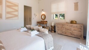 Une chambre spacieuse avec un grand lit, une commode en bois, une coiffeuse avec un miroir et une fenêtre donnant sur un paysage verdoyant.